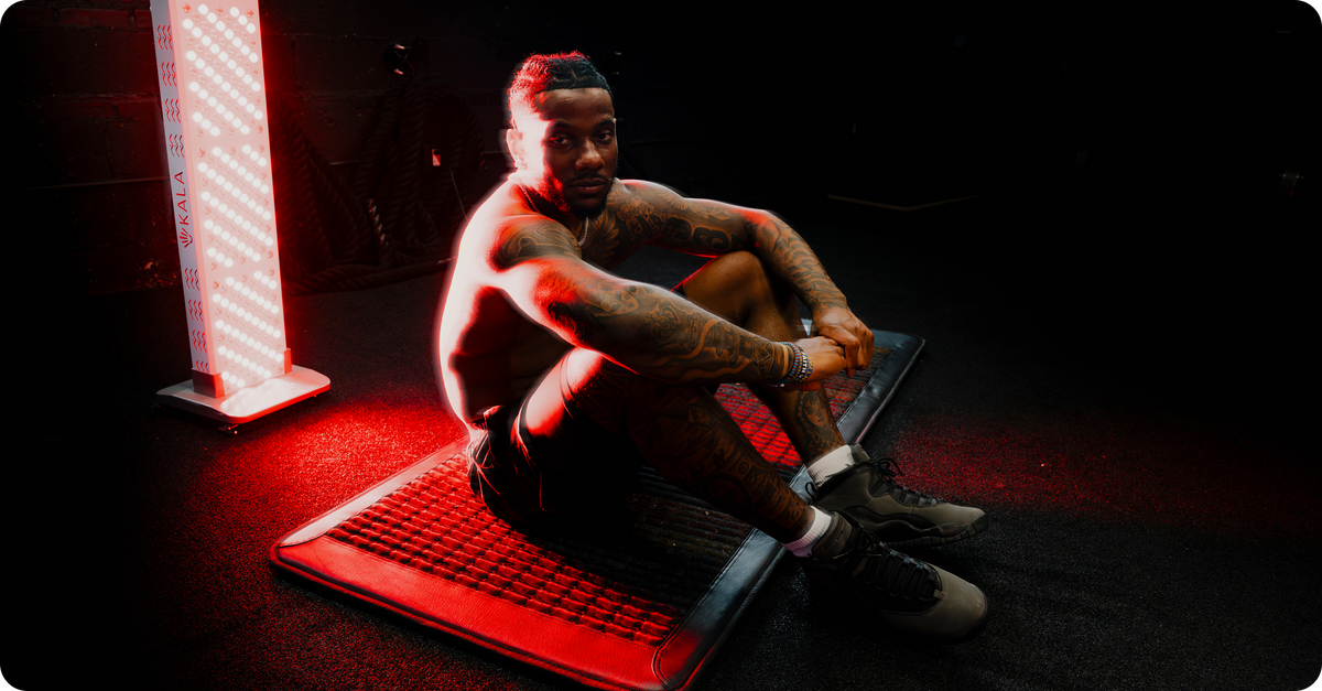 A tattooed man sits on a red LED light therapy mat in a dark gym environment, illuminated by a tall red light panel on the left.