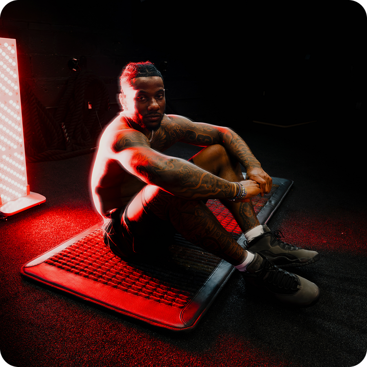 A tattooed man sits shirtless on a black mat in a dim studio setting, illuminated by dramatic red lighting. He is wearing dark shorts, white socks, and gray athletic shoes.