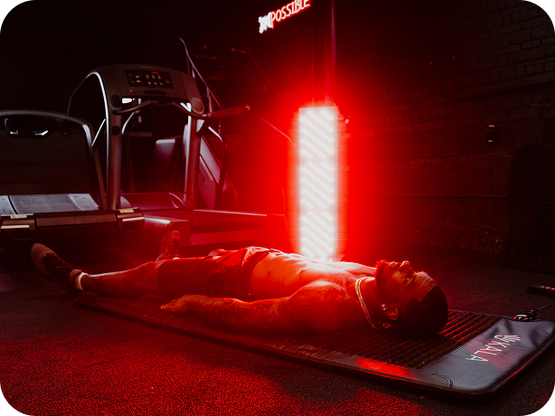 kala red light a person lies on a red light therapy mat in a dark gym setting, with a bright red light panel shining above them. A treadmill is visible in the background, and the brand name 'IMPOSSIBLE' appears on the mat and as a sign.