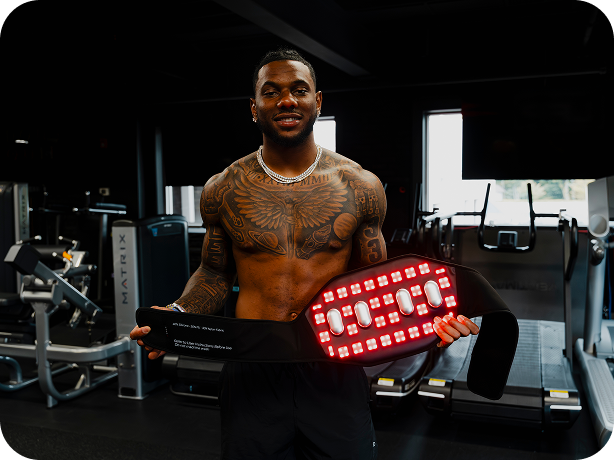 A shirtless tattooed man in a gym holds a red LED light therapy panel attached to a black strap. The background shows gym equipment and treadmills in a dark fitness center.