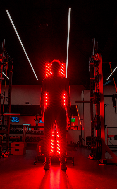 Silhouette of an athlete standing directly in front of a glowing, full-body Kala red light therapy panel setup inside a dark gym with overhead strip lighting.
