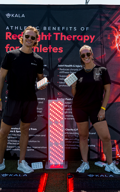 Two smiling fitness representatives in sunglasses holding handheld devices in front of a Kala athletic benefits banner, with a glowing Kala red light therapy panel standing between them.