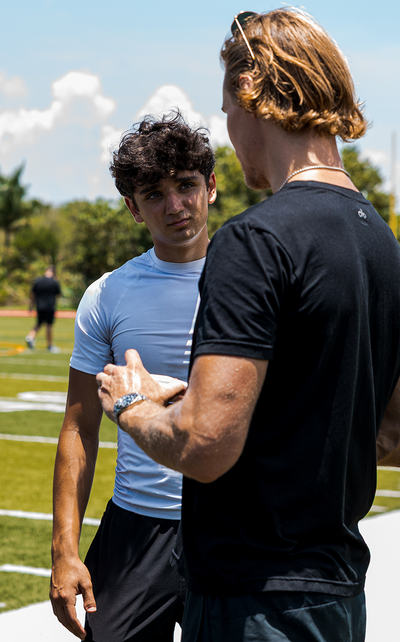 Two athletes discussing sports performance and recovery protocols on an outdoor football field, featuring a close-up profile of a trainer collaborating on Kala red light therapy integration.