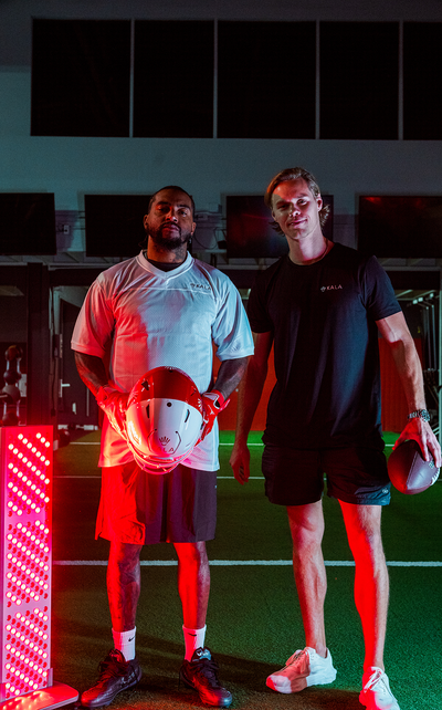 Professional football player DeSean Jackson holding a helmet alongside a trainer holding a football, standing next to a glowing Kala red light therapy panel on an indoor turf field.