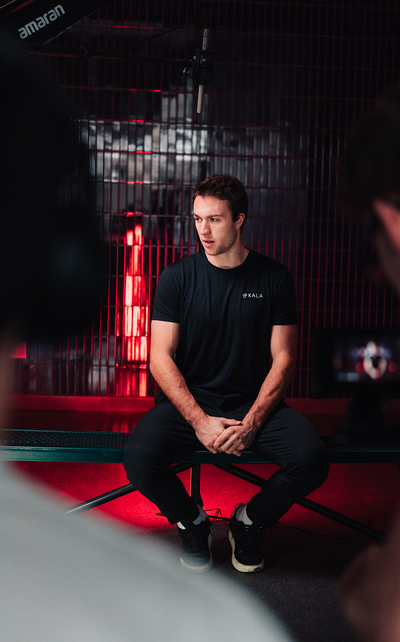 A man wearing a black Kala t-shirt sitting on a bench during a video shoot, with a glowing red background from a Kala red light therapy setup.