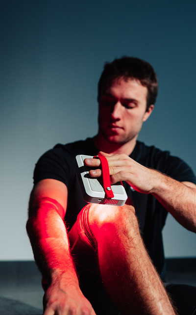 A man using a portable, handheld Kala red light therapy device with a red strap to target joint recovery on his knee.