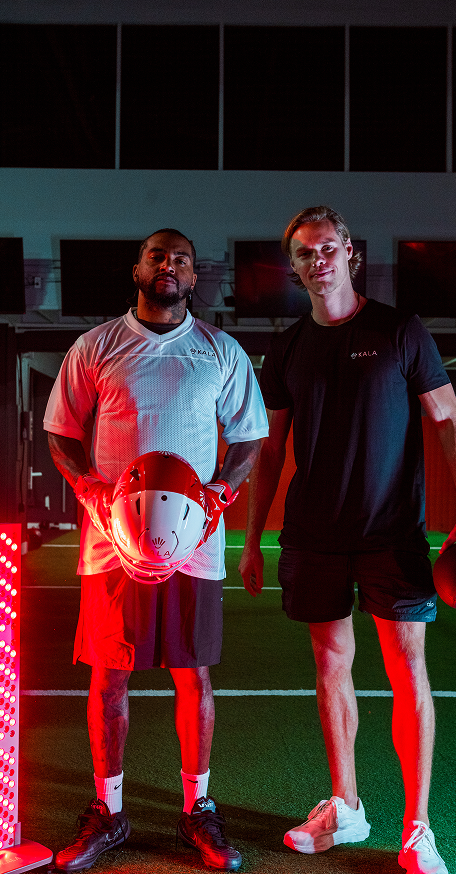 Football athlete standing with a trainer next to a Kala red light therapy panel inside a sports training facility.