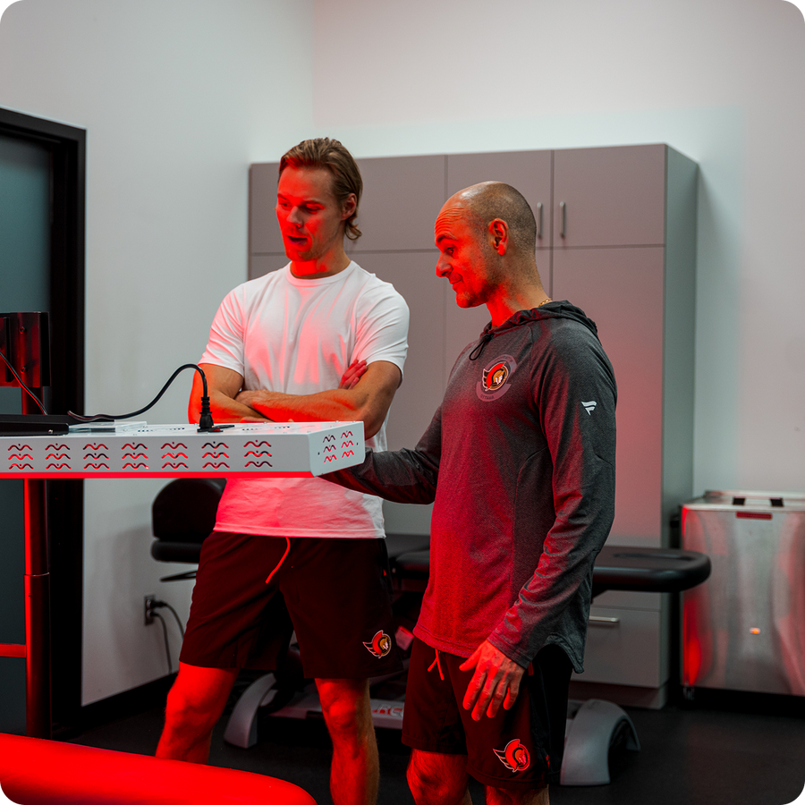 Two men in a sports wellness clinic examining a Kala red light therapy device on a table, next to an FAQ section about the Kala B2B Partner Program