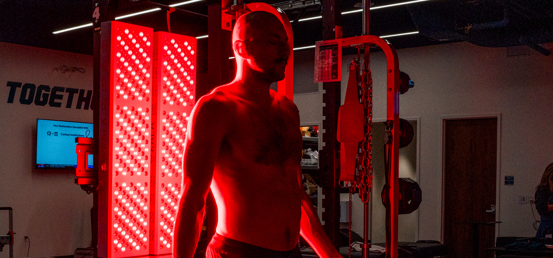 An athlete standing in front of large, glowing Kala red light therapy panels inside a professional sports training facility and gym.