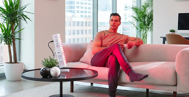 A man sitting on a white sofa in a bright corporate office lounge with large windows, utilizing a tabletop Kala red light therapy panel for workplace wellness.