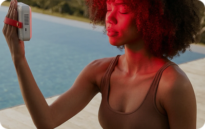 A woman outdoors by a swimming pool holds a small white and red device against her face. The scene is lit with strong red light, and she wears a ribbed brown tank top.