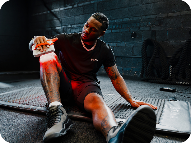 A man in a black athletic t-shirt and shorts sits on a mat in a gym, holding a white device that casts a red light onto his leg.