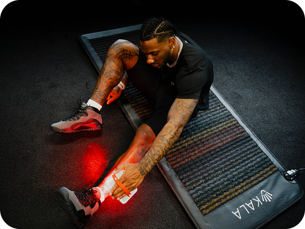 A man with tattoos sits on a dark textured mat on a black floor, applying a glowing red heat pack to his lower leg. The mat has the brand name 'YUKALA' printed on the corner.