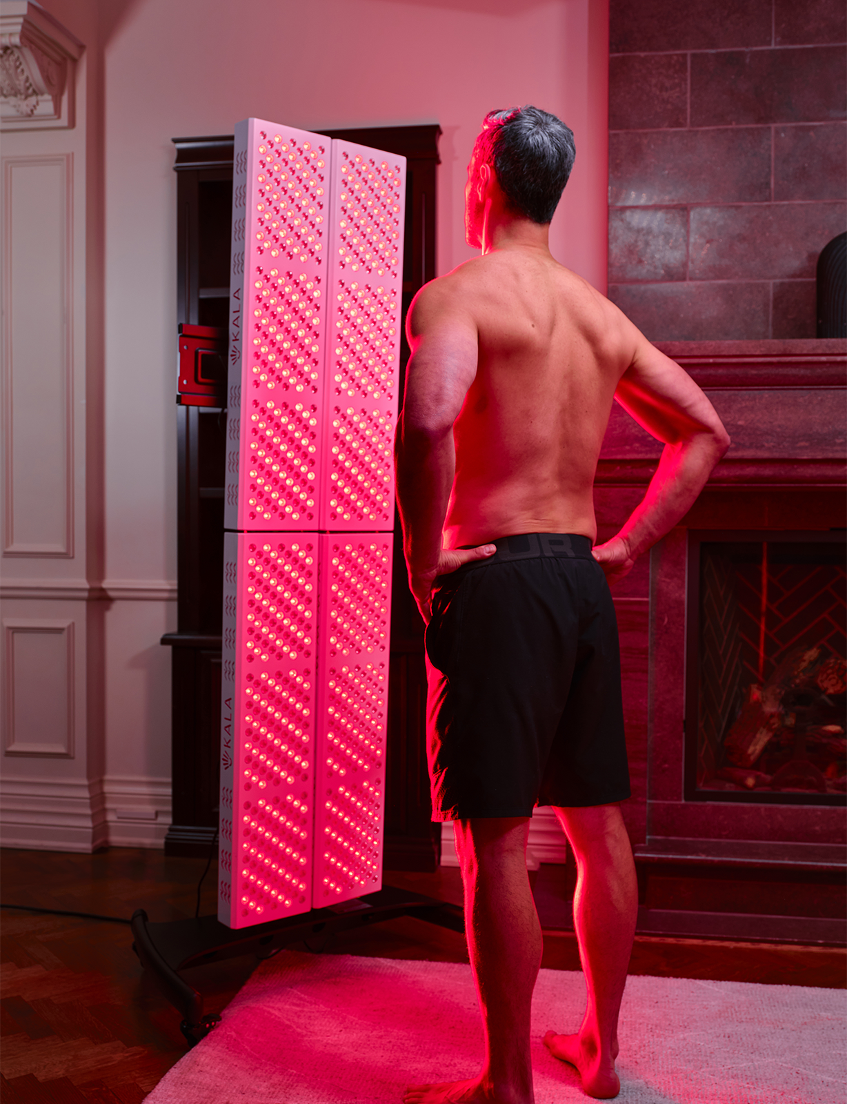 Man standing in front of a kala red light therapy device in a room with a fireplace.