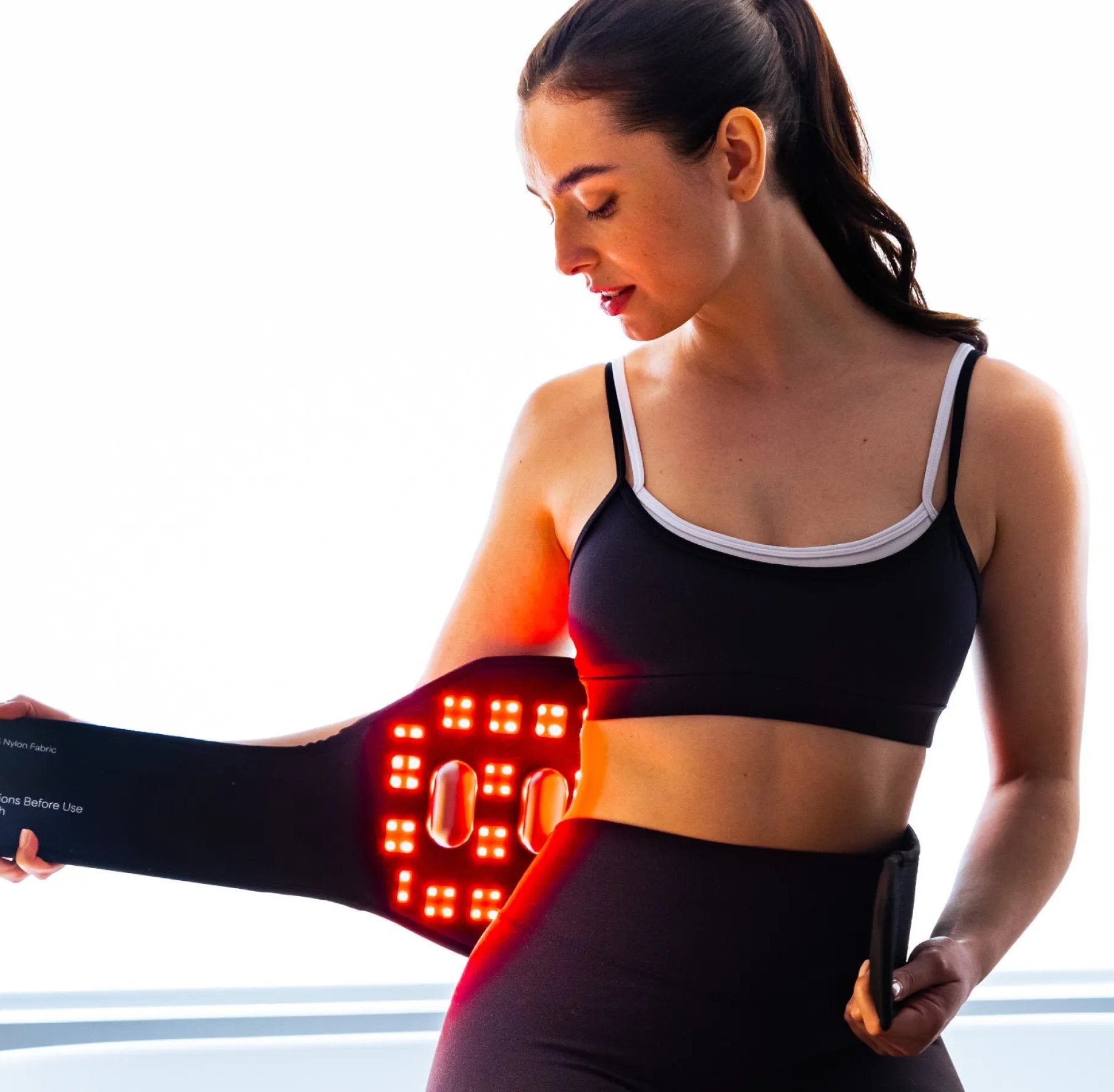 Woman in a bright bathroom holding a glowing red light therapy wrap to her waist.